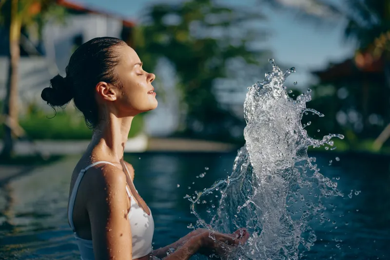 Smiling woman in a white swimsuit enjoys a pool splash, sun on her skin, water spraying around her, an outdoor wellness moment highlighting relaxation, lifestyle, and leisure in bright daylight.