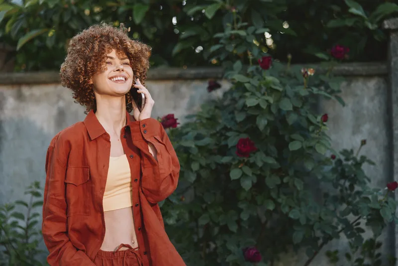 Confident woman with curly hair outdoors, wearing a red jacket and peach top, enjoying a sunny day in a garden with blooming roses, relaxed pose and natural style