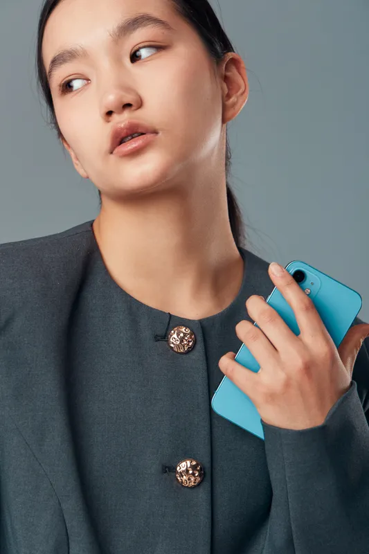 Young woman holding smartphone with blue case, looking sideways. Fashionable person wearing dark blazer with decorative buttons, studio portrait with soft lighting on gray background.
