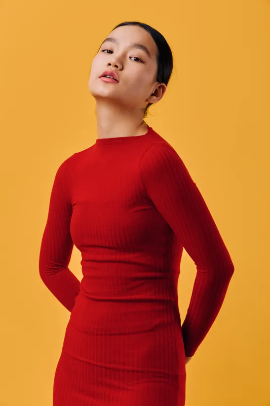 Young woman in red long sleeve dress posing confidently against yellow background with hands behind back. Portrait, fashion, style, and beauty with elegant look and clean composition.