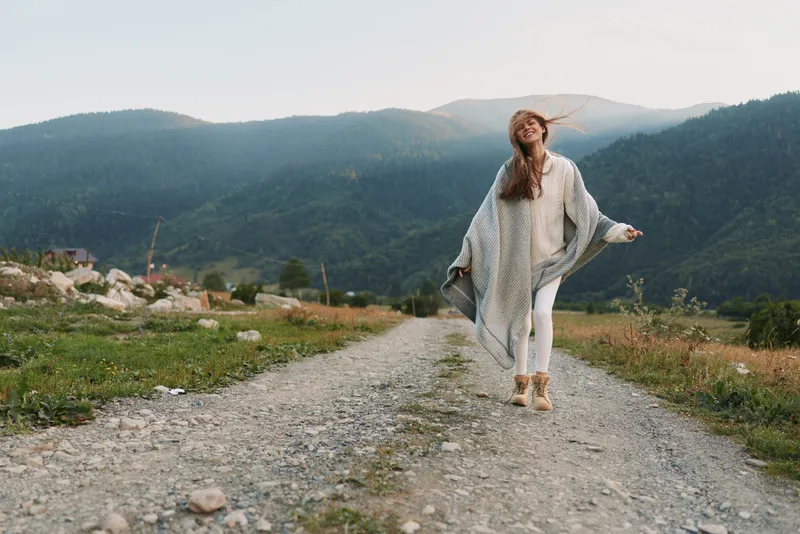 A young woman walks along a gravel road through a mountain landscape, wearing a cream poncho and ankle boots, savoring a carefree outdoor moment in wide open surroundings.