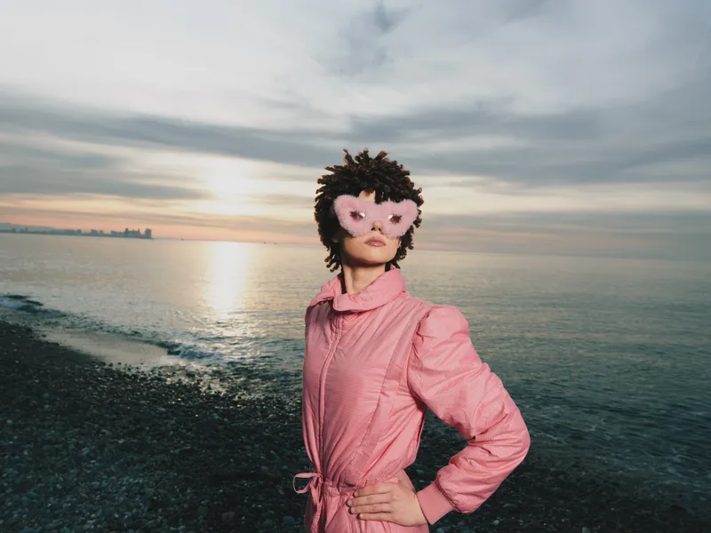 Woman on a beach at sunset wearing a pink coat and a masquerade mask, striking a confident pose as waves lap the shore and a colorful sky frames the scene.