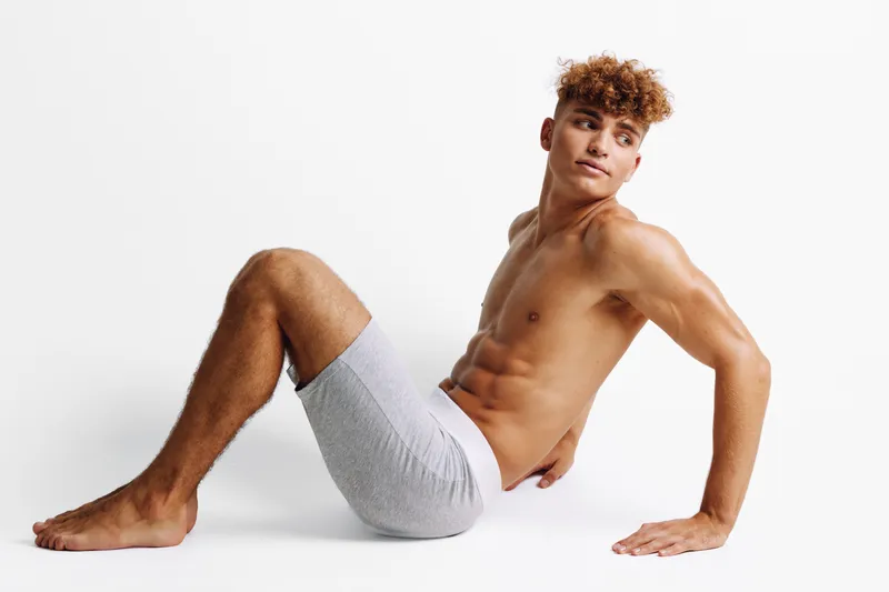 Young man with curly hair and muscular body posing shirtless in gray shorts on white background. Fitness, healthy lifestyle and male model with toned physique and confident expression.