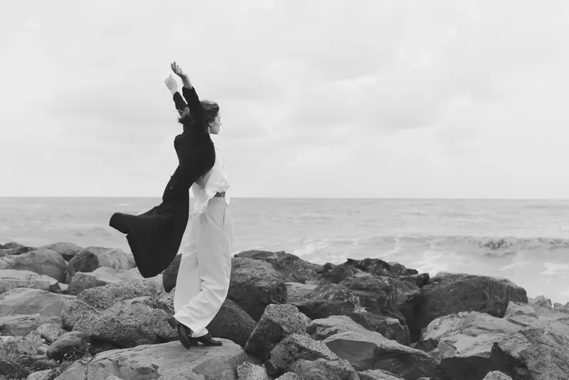 Woman on rocky shoreline raises arms in flowing dress, windblown silhouette, dramatic ocean backdrop, movement and freedom captured in a poetic coastal moment