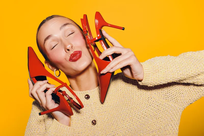 Young woman with closed eyes holds red high heel shoes near face, wearing beige sweater on bright yellow background. Concept of fashion, style, and shoe shopping.