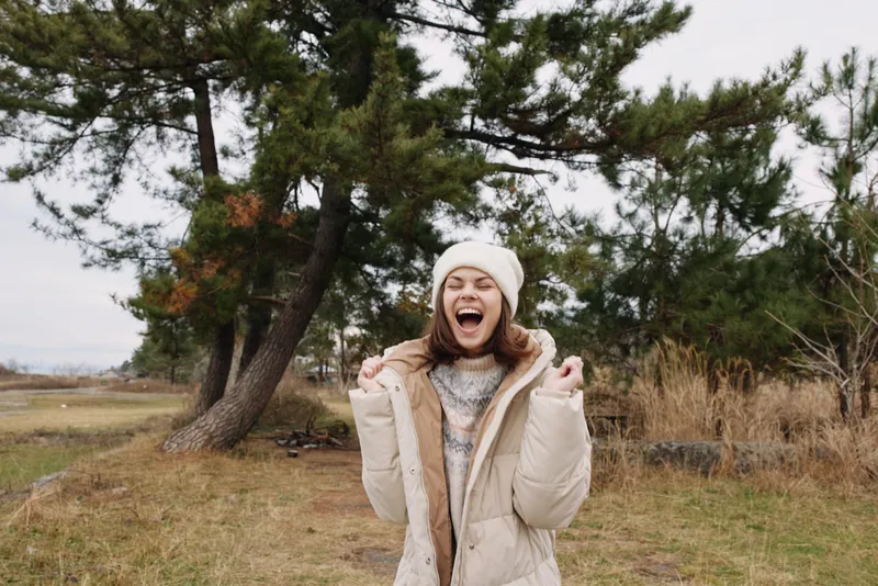 Outdoor woman in a padded coat and beanie celebrates with a bright smile in a windy field, trees in the background, capturing a mood of freedom and seasonal outdoor adventure.