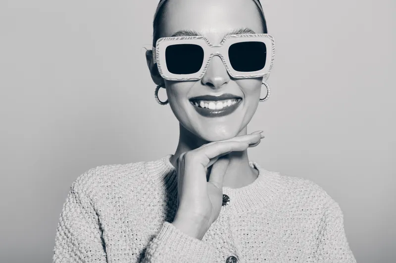 Smiling white woman with pale skin wears large sunglasses and knitted sweater posing with hand near chin in studio. Fashion and style concept.
