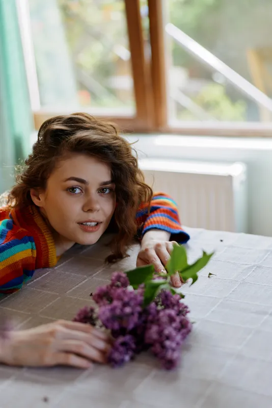 Cheerful woman at home, indoors, lifestyle moment with flowers on a table near a sunlit window, relaxed pose, soft colors, natural glow and calm atmosphere