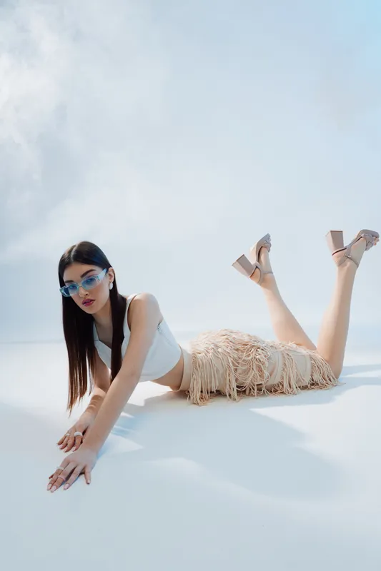 Young woman in stylish sunglasses and fringe skirt poses on white floor. She wears a white top and heels. Concept of woman beauty fashion and modern style in studio lighting.