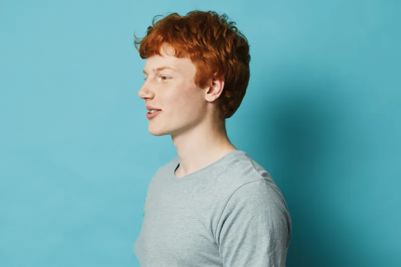 Young redhead man in gray shirt looking sideways on blue background, portrait with natural light and casual clothing for lifestyle and profile concept.