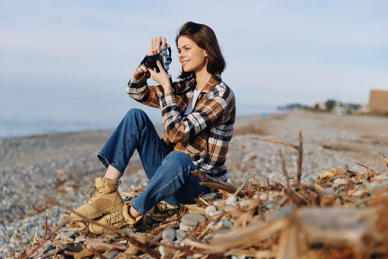 Photographer on a beach outdoor shoot, capturing candid moments with a camera among pebbles and leaves, wearing a plaid shirt and jeans, serene sea backdrop.