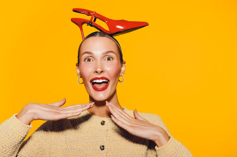 Happy woman with red high heel shoes on her head smiles widely on bright orange background. Concept of fun, fashion, creativity, and joyful expression in studio lighting.