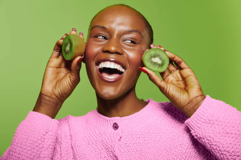 Happy African woman with dark skin holds two kiwi halves near face smiling broadly. Bright green background and pink sweater create vibrant contrast. Concept of healthy lifestyle and joy.