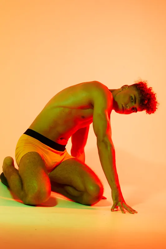 Young man with curly hair posing shirtless in yellow shorts on knees against orange background with dramatic red and green lighting for artistic and fashion photography concept.