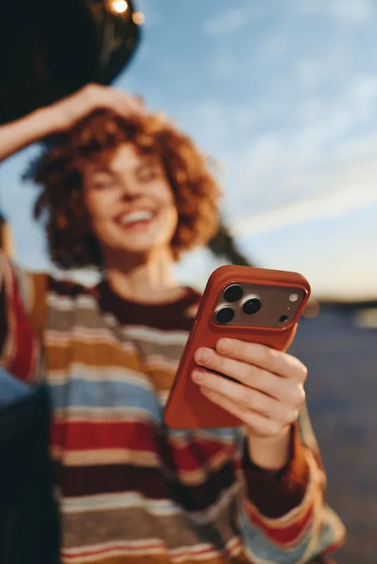 Woman lifestyle outdoor selfie moment, holding smartphone in hands, friends in background blur, warm sunset sky, casual striped sweater, candid portrait with smile, relaxed travel mood