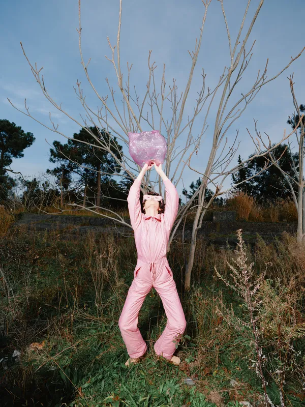 Person in a pink suit leaps outdoors, holding a pink heart-shaped fluffy prop above head, amid bare trees and open field under a clear sky, conveying joy and playful celebration.