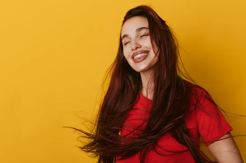 Woman with long hair wearing red shirt smiling with closed eyes on yellow background. Portrait of happy female enjoying moment with natural expression and casual style indoors.