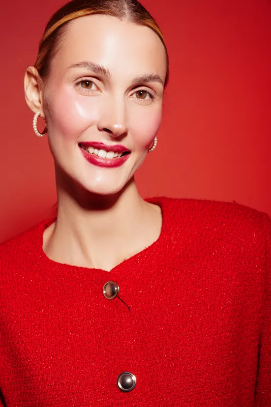 Smiling woman with red lipstick and red sweater poses against red background. Close-up portrait with hoop earrings and natural makeup. Concept of fashion, beauty, and confidence.