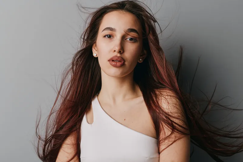 Woman with long hair and natural makeup wearing white top posing against gray background. Female model with serious expression and loose hair in studio portrait with soft lighting.