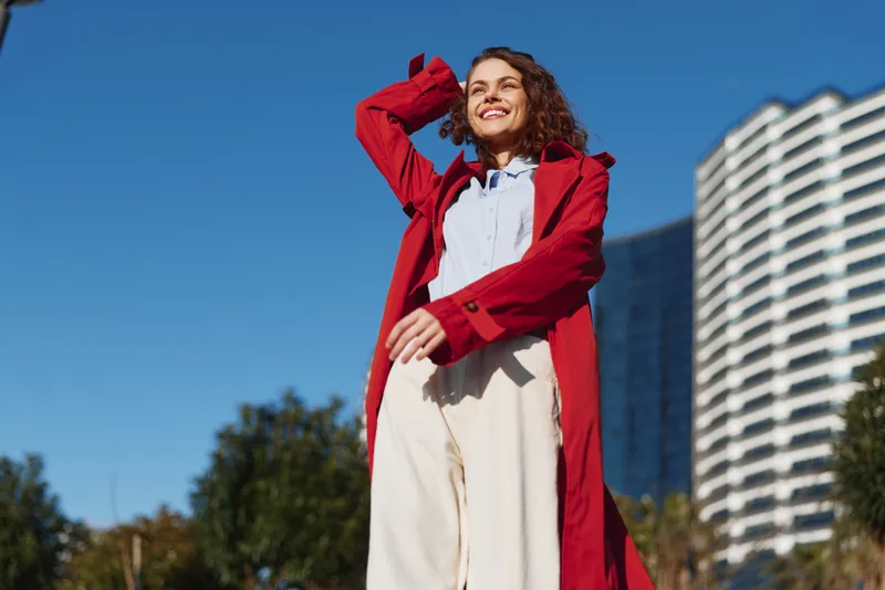 Smiling woman in red coat walking outdoors on sunny day with blue sky and modern building background, casual style, natural light, confident expression