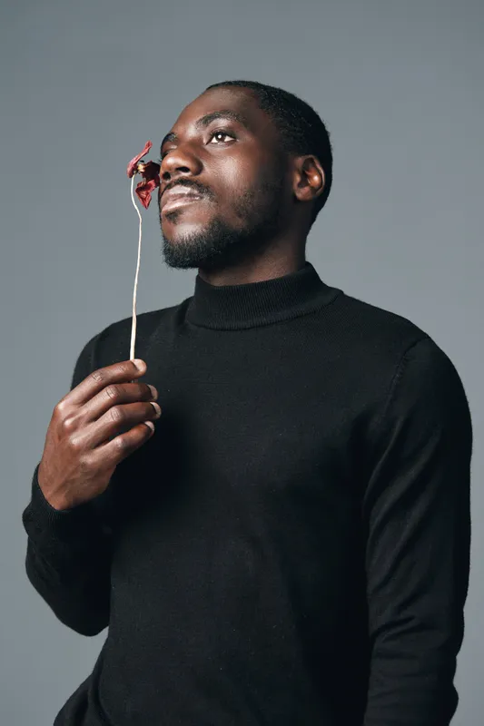 African man in black turtleneck holding dried flower near face in creative conceptual visual style with thoughtful expression on gray background in studio portrait.