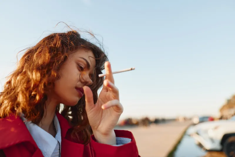 Young woman outdoors, smoking a cigarette in a red coat, sunlit moment, wind in hair, casual pose, natural tones, urban backdrop