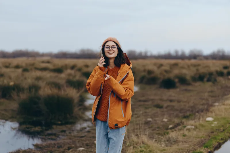 Woman lifestyle in outdoors autumn field, smiling with orange hooded jacket and glasses, standing on path near water puddle, casual nature portrait with calm mood in fresh air daylight