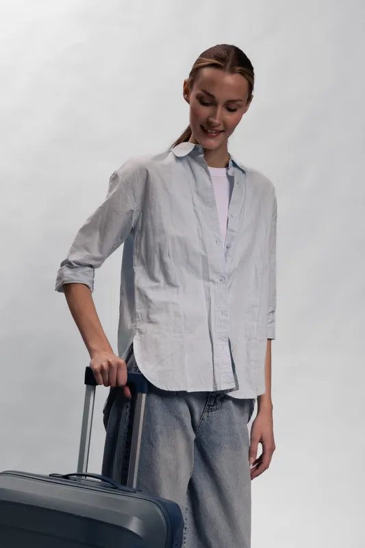 Young woman in casual shirt and pants stands with suitcase, looking down with slight smile. Concept for travel, vacation, leisure, and casual fashion lifestyle in neutral studio setting.
