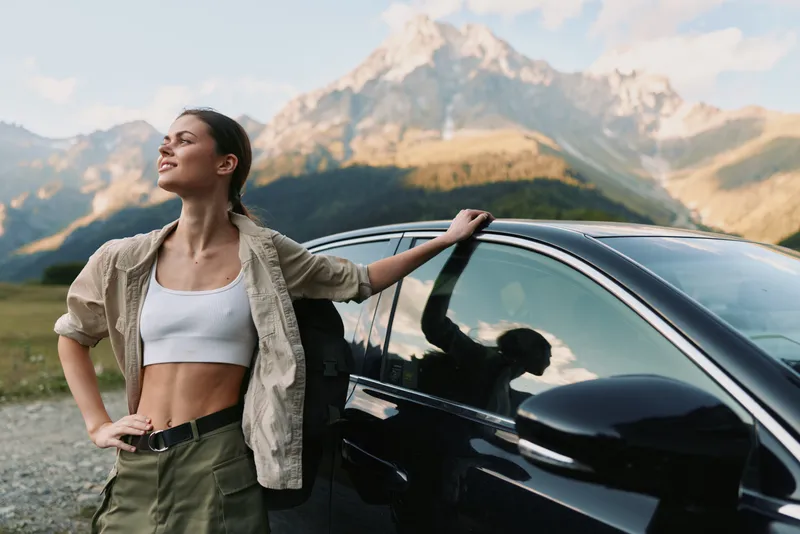 Confident woman leaning against a sleek car in a sunlit mountain landscape, capturing a carefree outdoor lifestyle moment with casual wardrobe and adventurous mood.