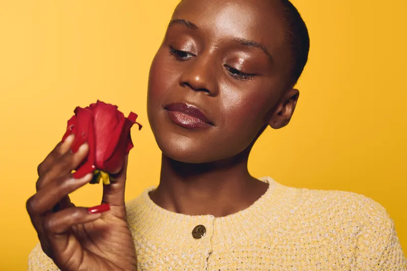 Close-up of a thoughtful dark skinned African woman holding a red rose against yellow background. Concept of beauty, skincare, self-care, and natural elegance with floral element.