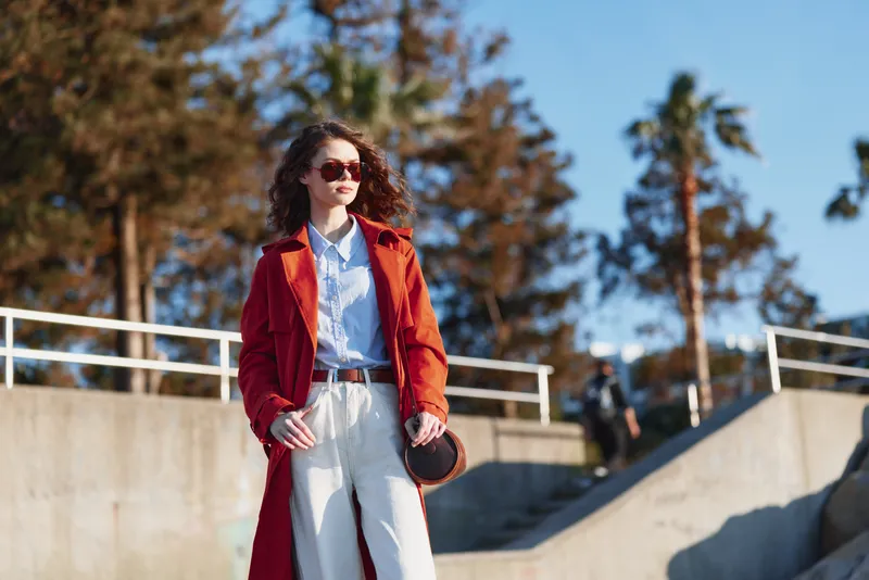 A stylish woman with curly hair in a red coat stands on sunlit stairs, sunglasses on, exuding bold confidence and street fashion in a modern urban setting.
