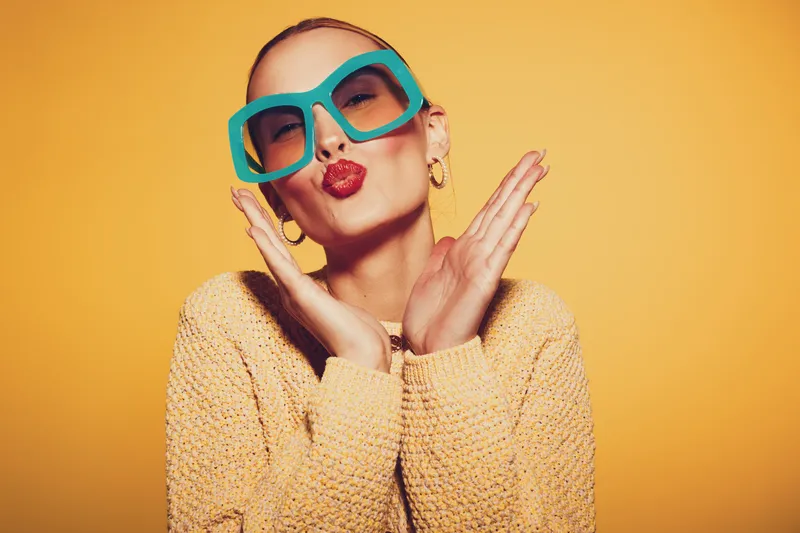 Young woman in oversized turquoise sunglasses and yellow sweater blows a kiss with hands near face on bright yellow background. Concept of fashion, beauty, and playful attitude.