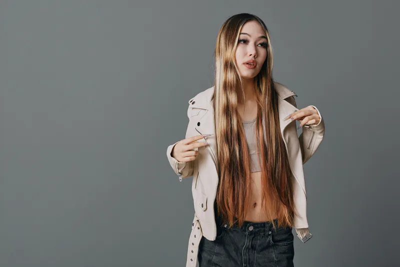 Young woman with long hair wearing light jacket and casual top posing with hands showing peace gesture on gray background. Fashion, style, confident, studio portrait.