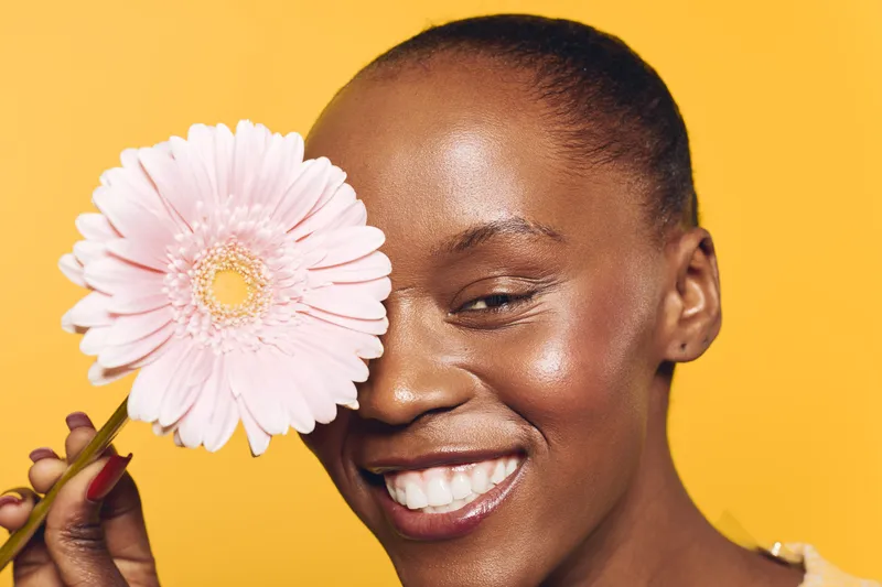Happy African American woman with dark skin holds pink flower near face smiling joyfully on yellow background. Concept of beauty, skincare, wellness, natural, and floral health care.