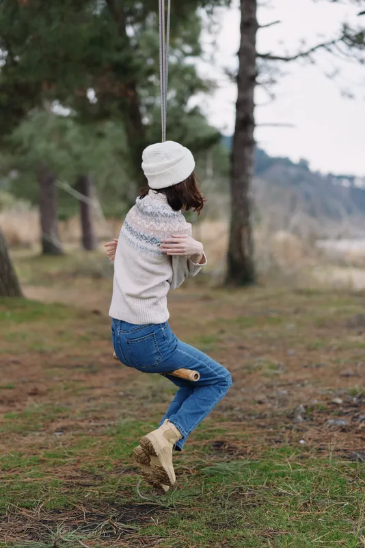 Person on a swing in a park, wearing a knitted sweater, beanie and boots, enjoying a calm outdoor moment among trees and open space.