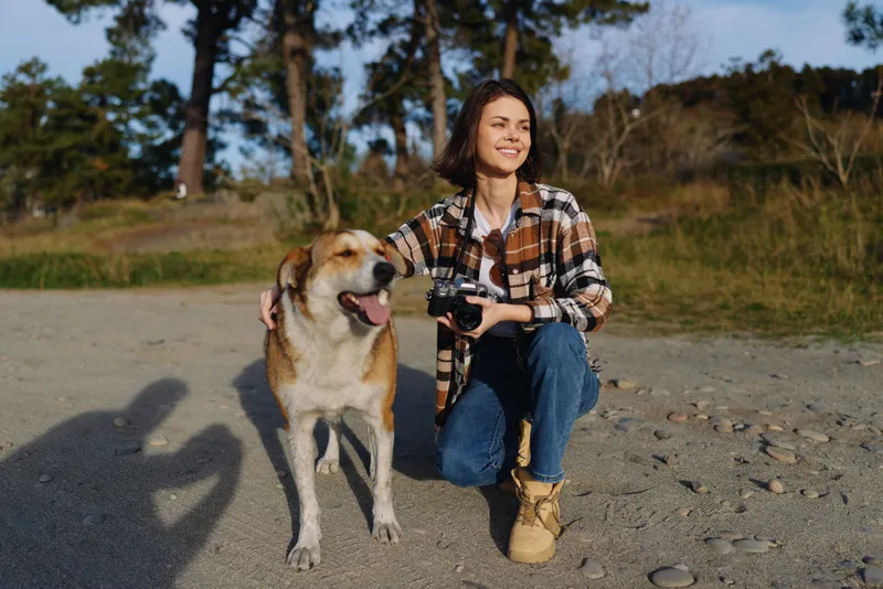 Woman photographer kneels beside a playful dog on a rocky beach, holding a camera as warm sunlight and open scenery frame an outdoor portrait full of joy and companionship