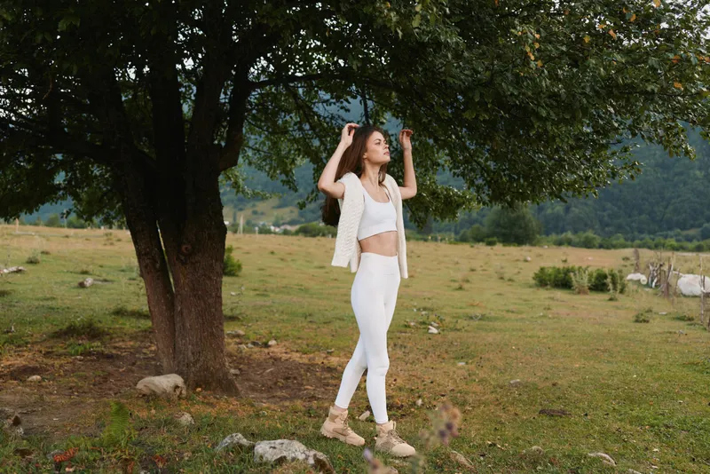 Relaxed woman outdoors in a white outfit under a leafy tree, stretching arms above head in a sunlit field, highlighting lifestyle, wellness, confidence, and nature everyday.