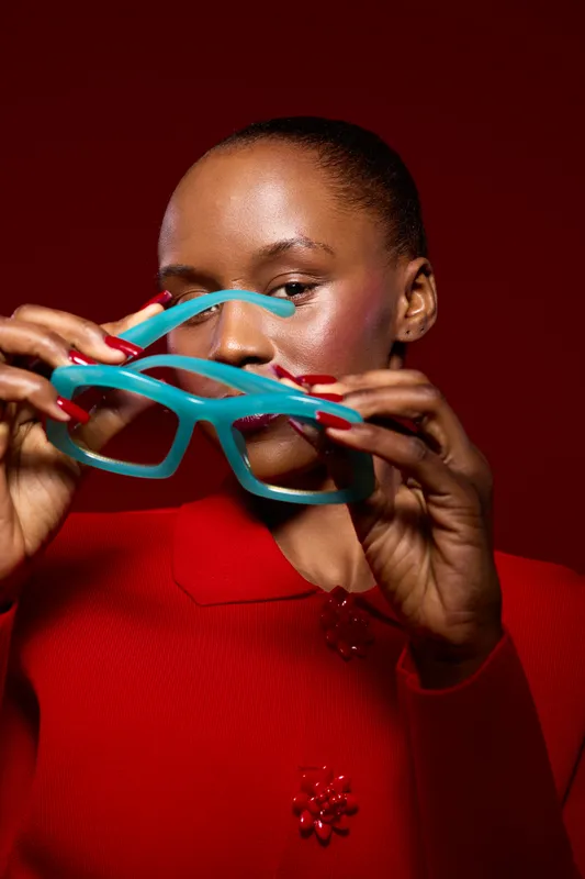 Close-up of a dark skinned African woman holding turquoise glasses in front of her face wearing a red outfit. Studio portrait with vibrant colors and fashion concept.