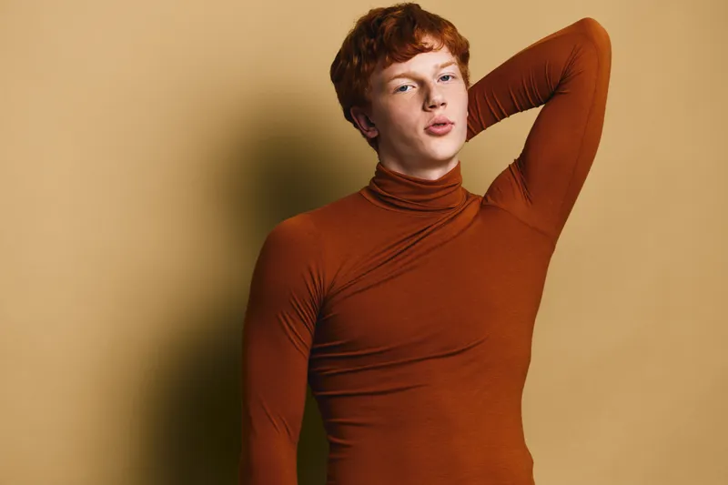 Portrait of young man with red hair in brown turtleneck posing with hand behind head on beige background. Closeup of confident male model with casual style and relaxed expression.