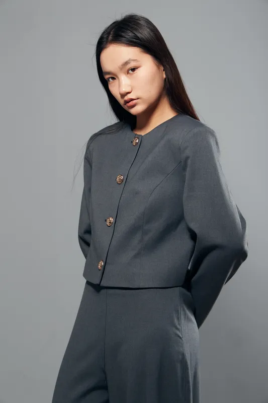 Young woman wearing gray knitted outfit with buttoned cardigan and matching pants posing with hands behind back on gray studio background. Fashion model with long hair in casual attire.