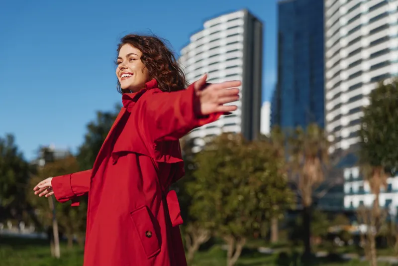 A smiling young woman in a red coat stands in a sunny urban park with arms open, expressing confidence and a free, adventurous mood.