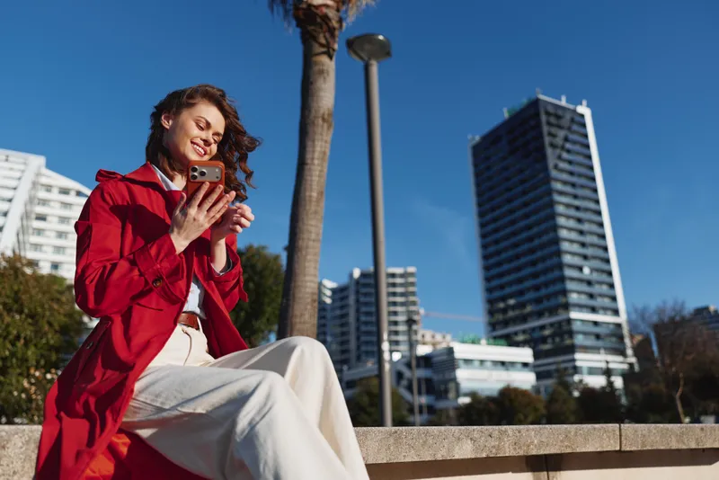 A smiling woman in a red coat sits on a low wall, looking at her phone. Urban buildings and palm trees frame a casual, modern lifestyle moment in the city.