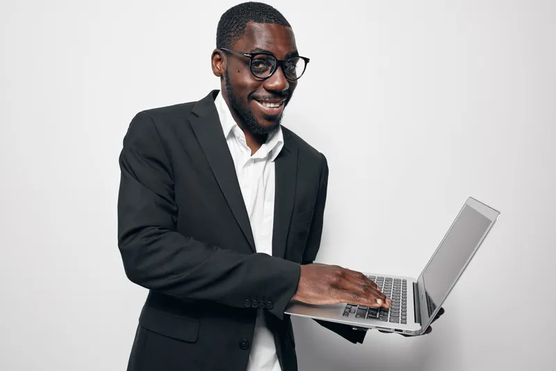 African American man with dark skin wearing glasses and black suit using laptop computer for work or communication on white background. Business professional smiling and typing on device.