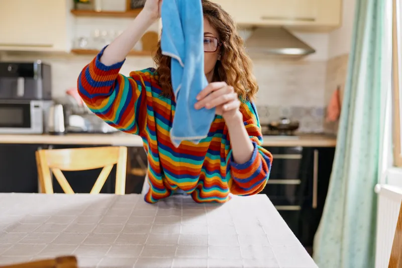 A woman in a striped sweater dries a blue cloth at a kitchen table, depicting a candid home cleaning moment. A realistic domestic scene focusing on lifestyle and everyday chores.