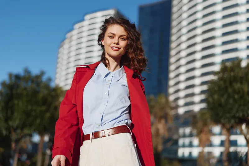 Smiling woman wearing a red coat and light blue shirt outdoors in an urban setting, with tall buildings in the background, casual fashion, confidence, and sunny mood.