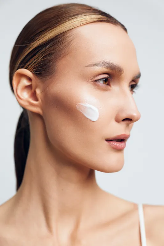 Young woman with clear skin applying white cream on cheek, natural skincare routine, smooth complexion, soft lighting, neutral background, beauty and wellness concept