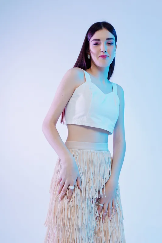 Young woman in stylish white crop top and fringed skirt poses confidently in soft blue studio lighting. Concept for woman beauty fashion, elegance, and modern style.