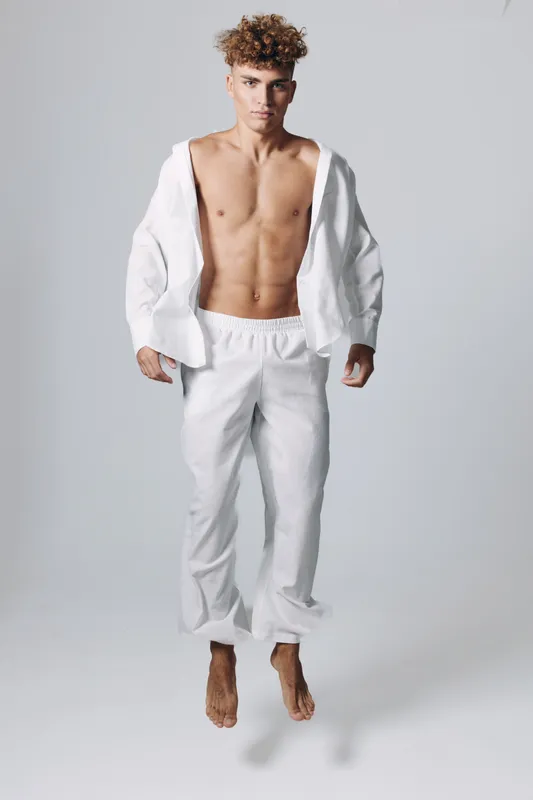 Young man with curly hair jumping in white loose pants and open shirt, bare chest visible, isolated on gray background with shadow below for fitness or fashion concept.