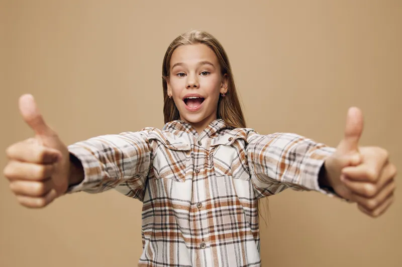 Girl with long hair smiling and showing thumbs up gesture wearing checkered shirt on beige background for success and approval concept.
