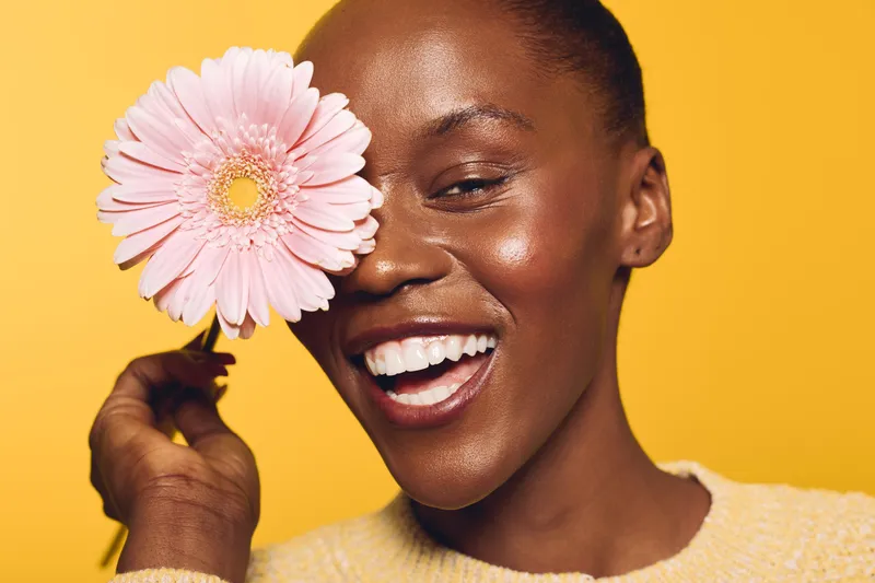 Smiling dark skinned African woman holds pink flower near eye on yellow background. Concept of beauty, happiness, skincare, natural glow, and positive emotions with floral element.